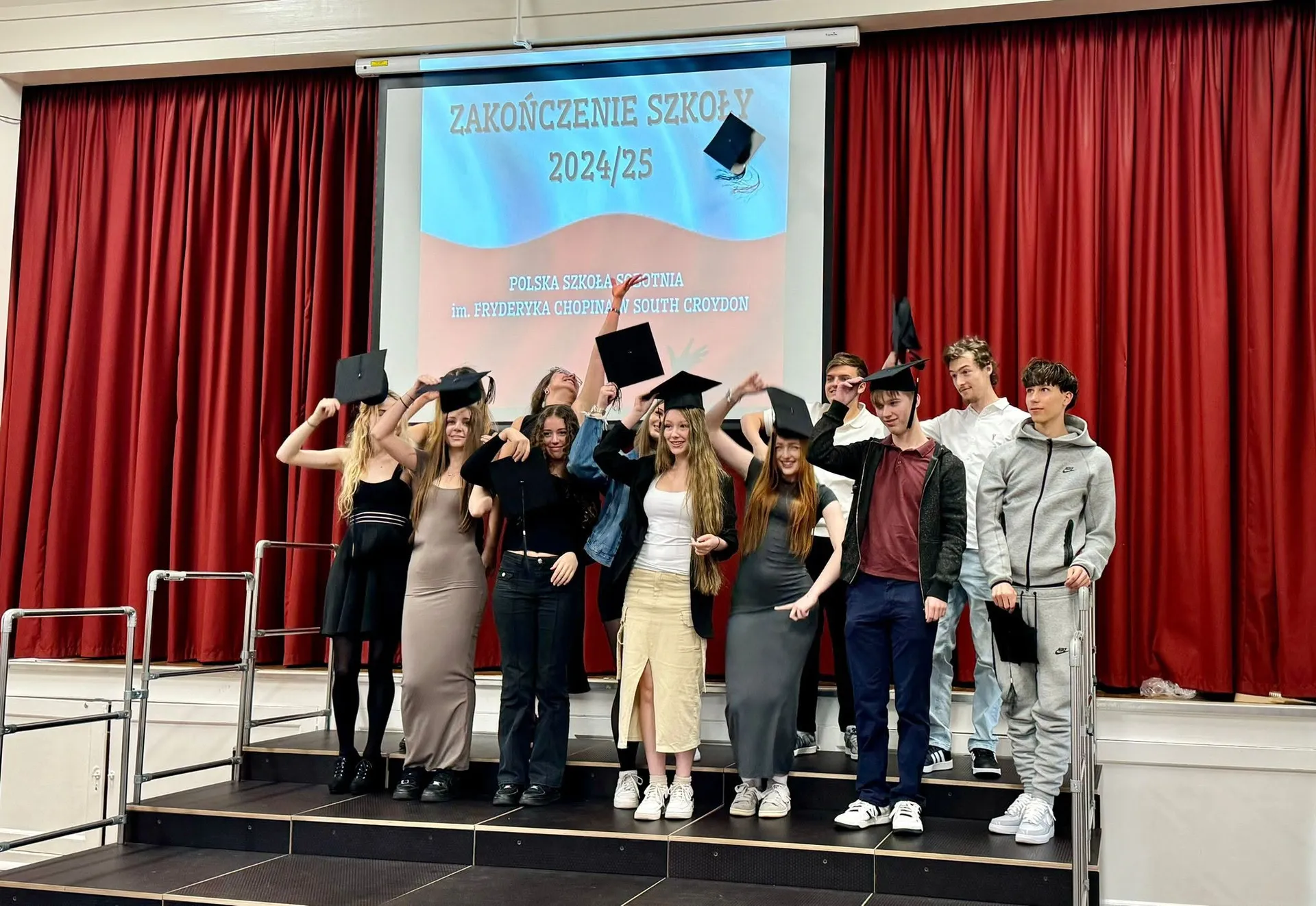 Klasa A-level pozuje na scenie pod ekranem „Zakończenie szkoły 2024/25”; kilka biretów unosi się w powietrzu.
