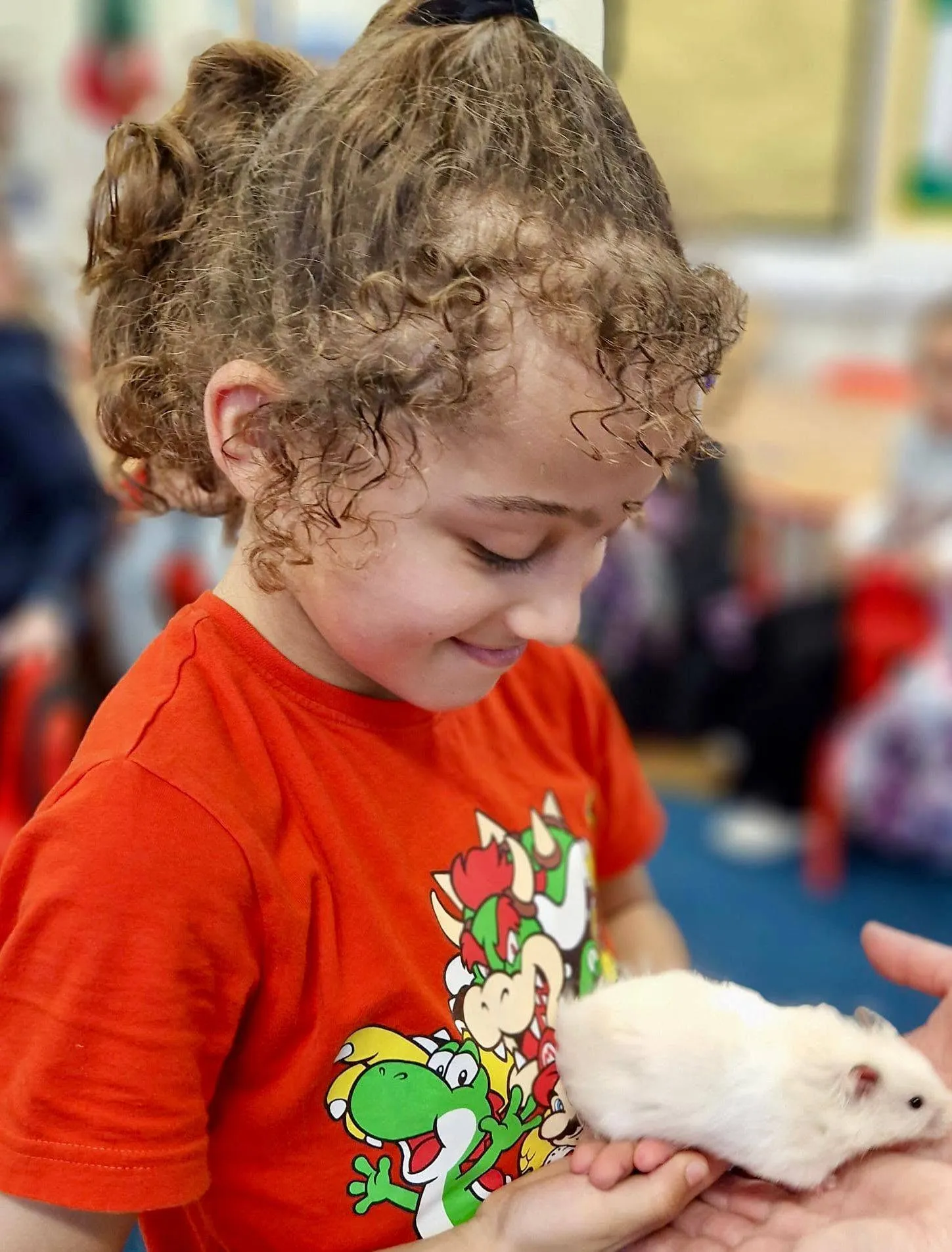 A smiling pupil looks at Coco, a white hamster, held on the teacher's hand.