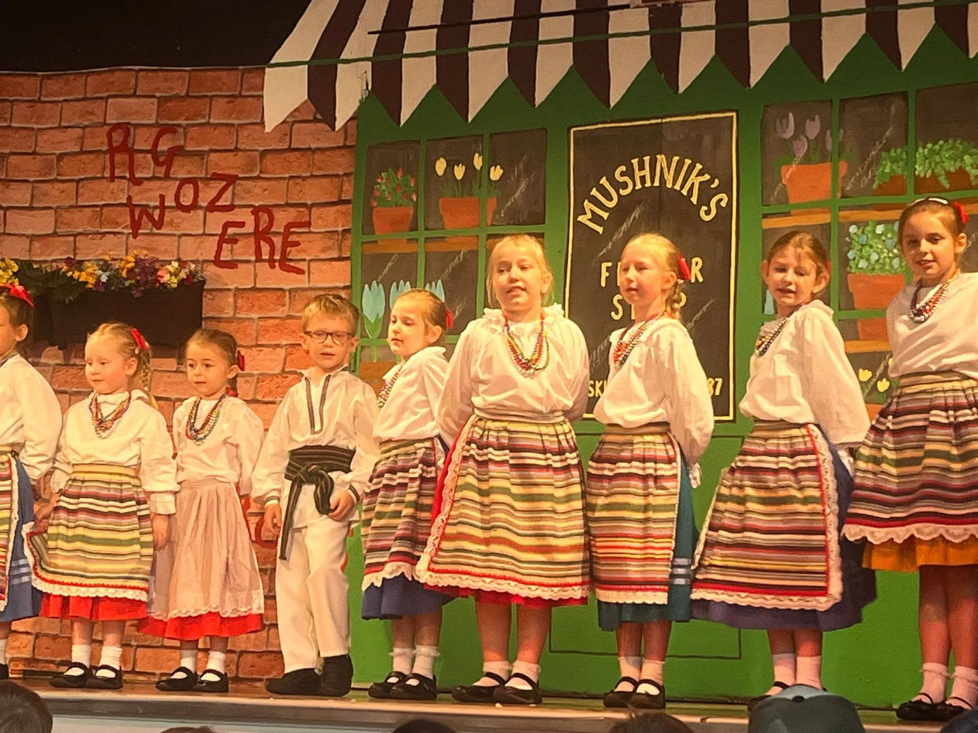 Dzieci z zespołu Karolinka w pasiastych spódnicach i białych strojach ludowych stoją w rzędzie na scenie.