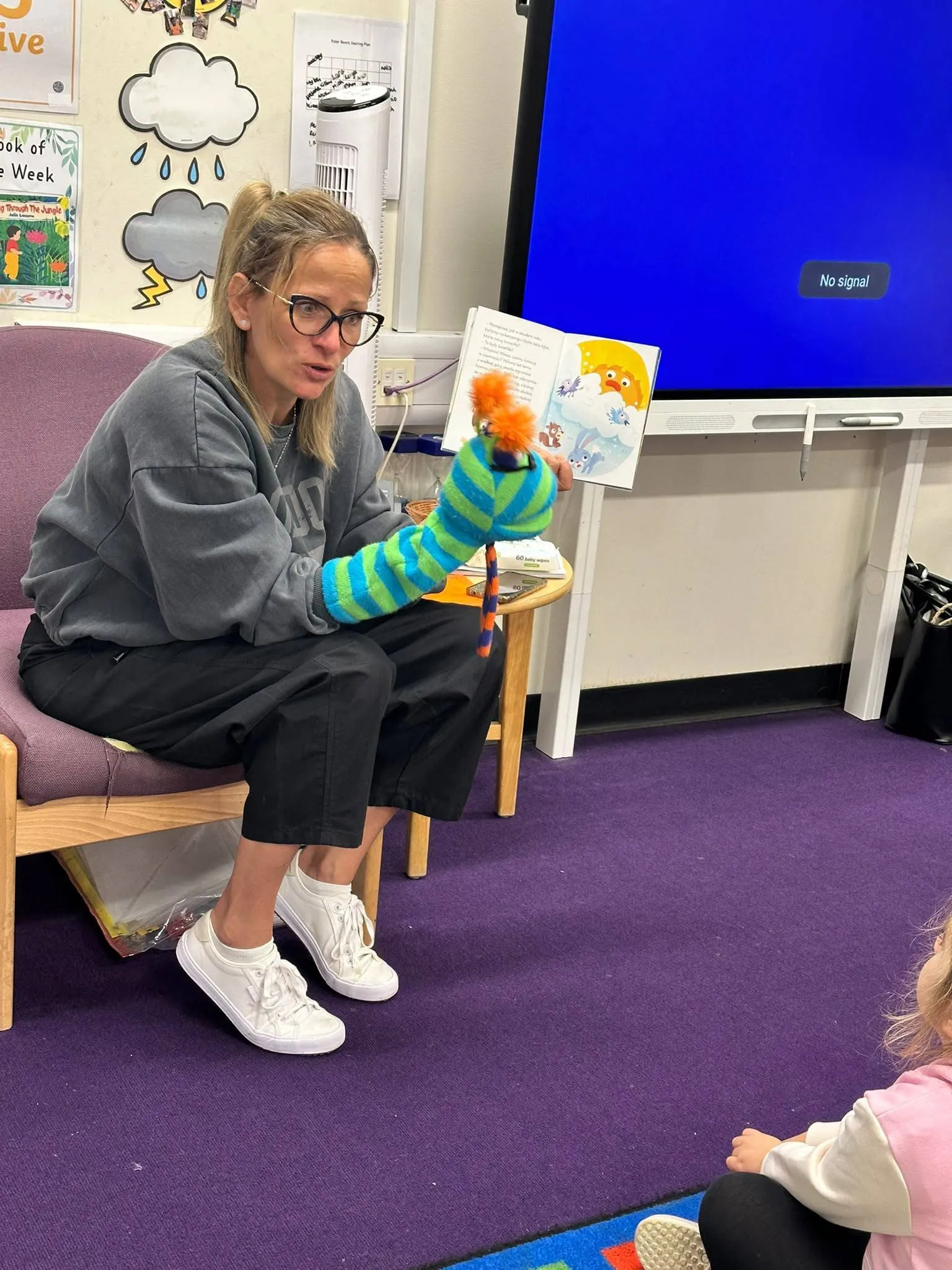 The teacher, sitting on a chair in the classroom with a sock puppet on her hand, reads a book to the children.