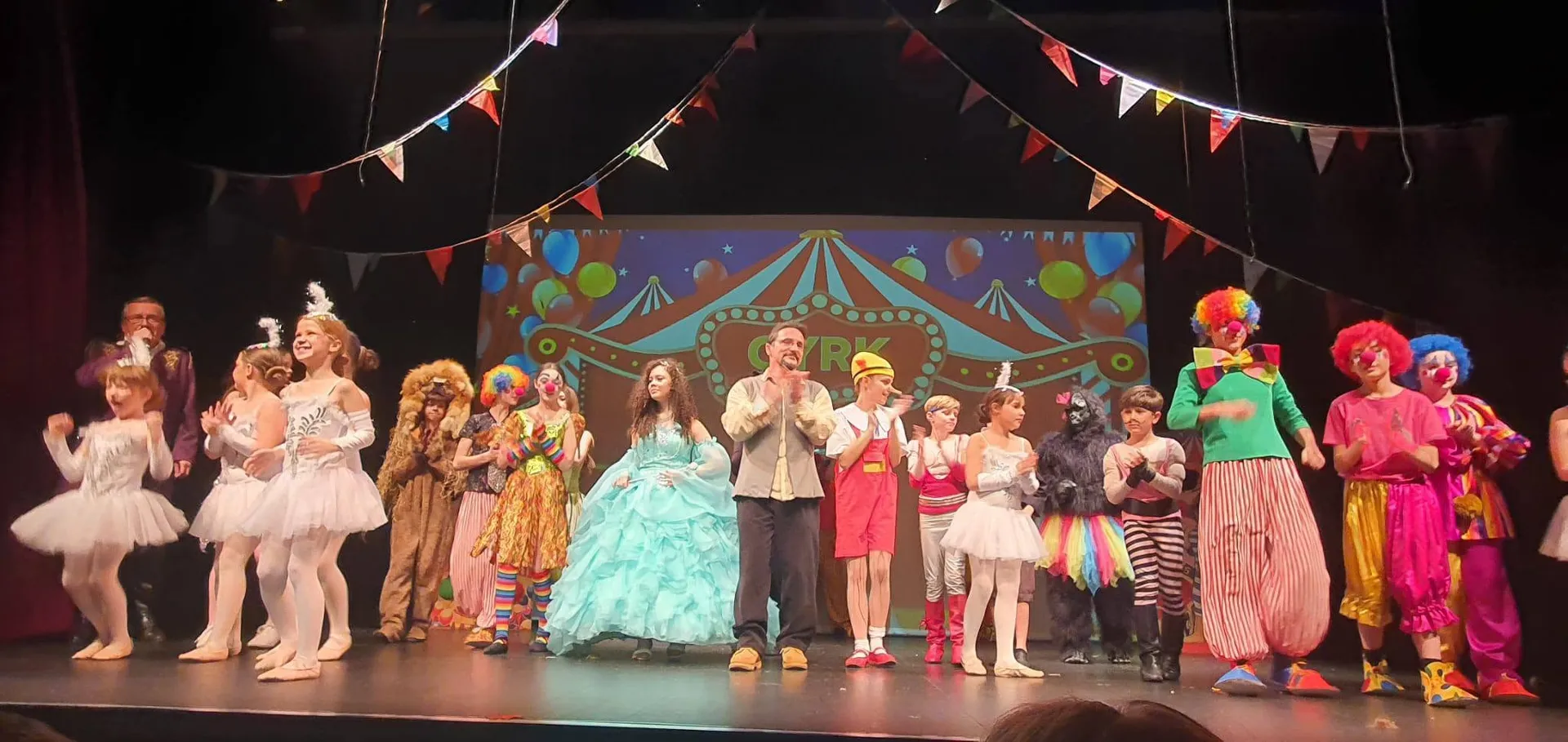 Obsada przedstawienia „Pinokio” Teatru Syrena w kolorowych kostiumach na scenie z cyrkową dekoracją.