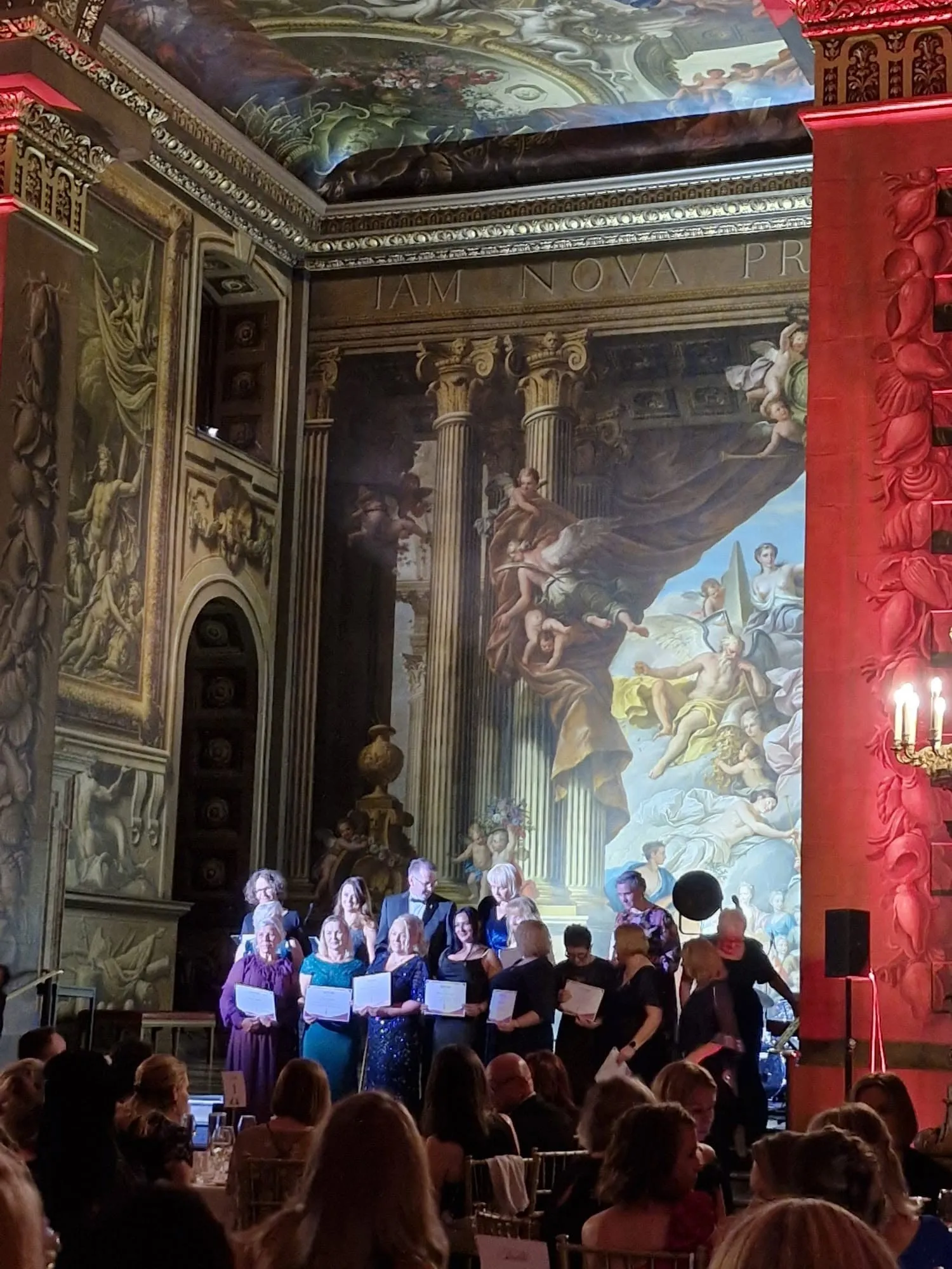 Widok Painted Hall z malowidłami na ścianach i sklepieniu; na scenie grupa wyróżnionych, przed nią goście gali.